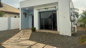 A modern building with a flat roof, a glass door entrance, and a small garden area with a pathway. - Beachside Studio with a Curated Library (P32W+99C Prampram)