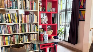 A room with a bookshelf filled with books, a red shelf unit with various items, a potted plant, and a window with curtains. - Beachside Studio with a Curated Library (P32W+99C Prampram)