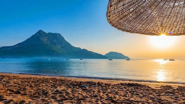 A beach with a chair and a woven umbrella, a mountain in the background, and boats on the water. - zeyos lodge (kumluca)