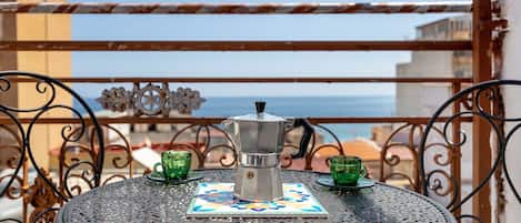 Una mesa redonda de metal con una cafetera y dos tazas verdes, ubicada en un balcón con vistas al mar.