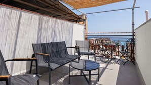 Un'area salotto esterna con tavolo e sedie, vista sul mare e una parte coperta con tetto di paglia.
