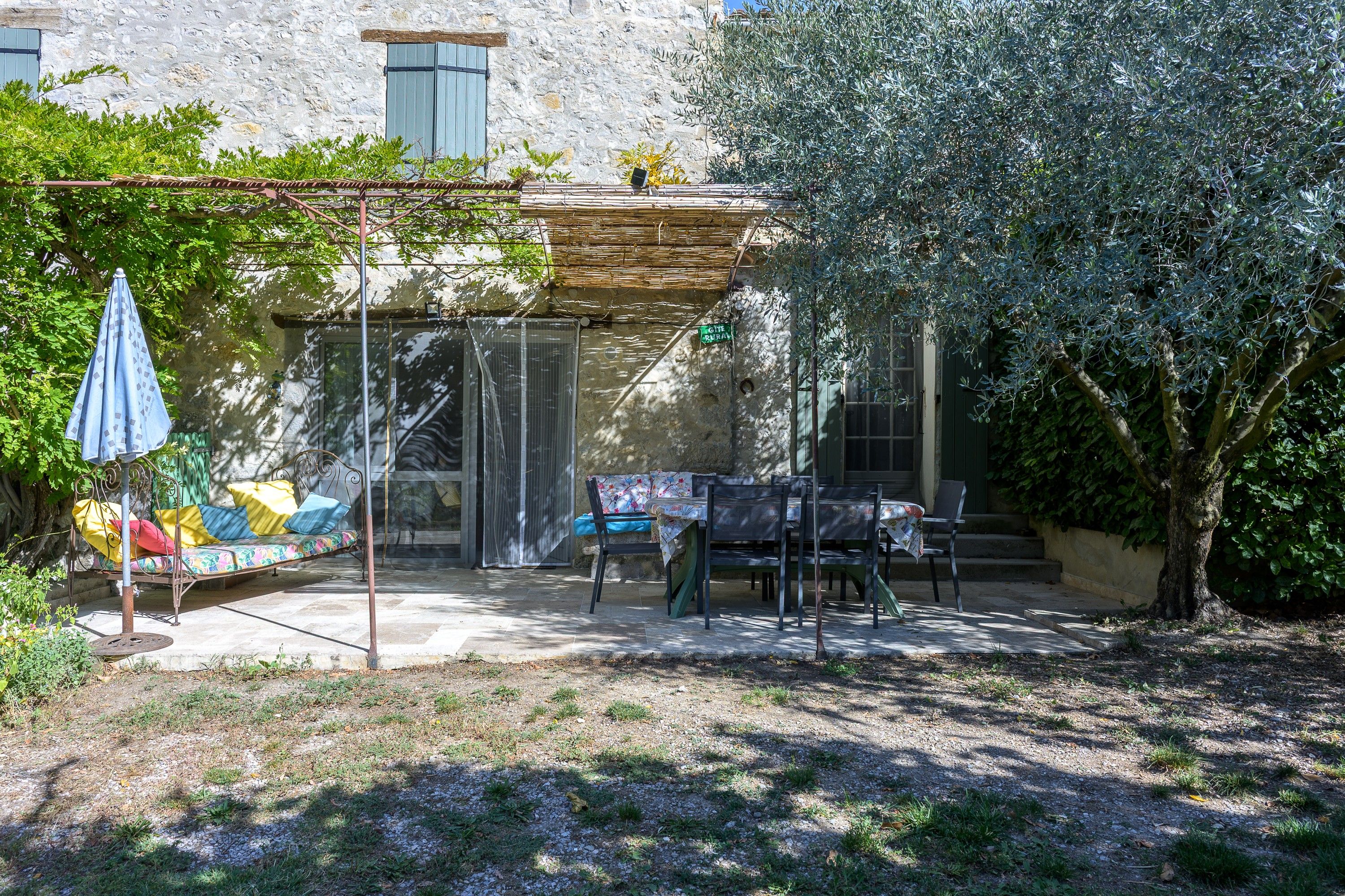 A rustic outdoor seating area with a metal table and chairs, a covered bench, and a pergola with a blue umbrella.