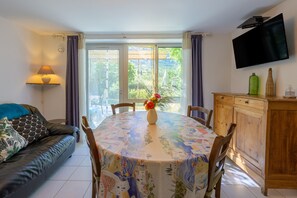 Ein Essbereich mit einem runden Tisch, der mit einer bunten Tischdecke bedeckt ist, eine Couch mit gemusterten Kissen, ein an der Wand montierter Fernseher und Blick ins Grüne durch die Fenster.