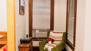 Quarto com poltrona verde, piso de madeira, ventilador de teto, radiador e janela com persianas.