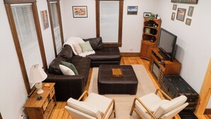 Uma sala de estar com um sofá de couro marrom, duas poltronas brancas, uma mesa de centro de madeira, uma televisão e uma lareira.
