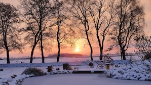 Schneebedeckte Landschaft mit Bäumen und einer zugefrorenen Wasserfläche im Abendlicht.