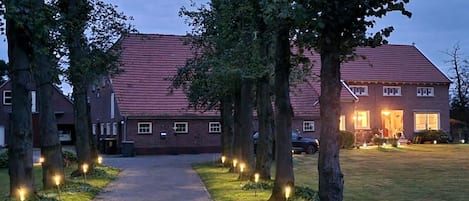 A pathway leading to a house with a red roof, surrounded by trees and lit by ground lights.