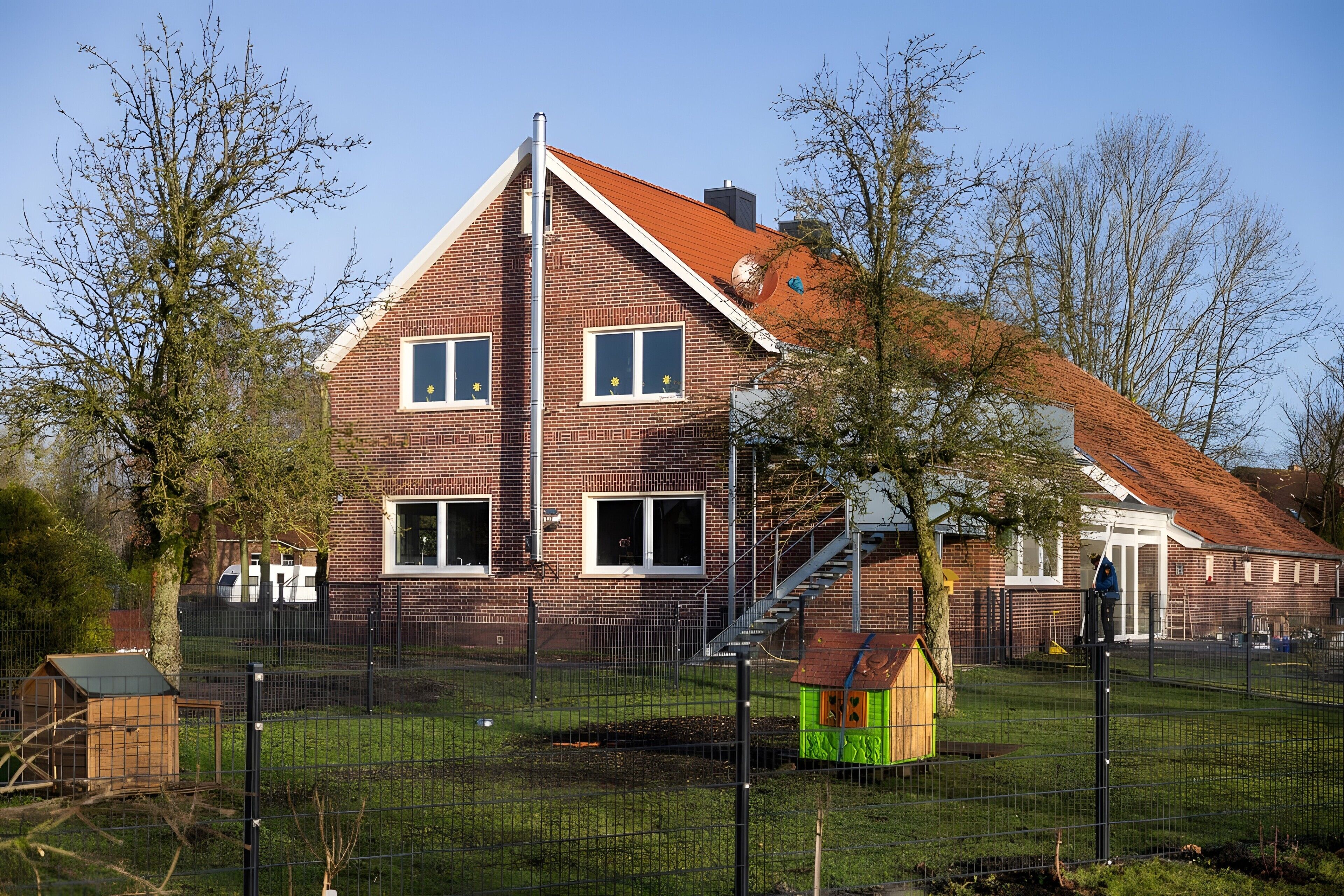 Ein zweistöckiges Backsteinhäuschen mit rotem Dach, einem sichtbaren Schornstein und einem umzäunten Garten mit einem kleinen, grünen Spielhaus.