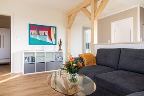 A living room with a dark grey sofa, a glass coffee table with a vase of flowers, a white shelving unit with books and glasses, and a colorful painting on the wall. - Apartment 'Großes Mehr' with Wi-Fi (Südbrookmerland)