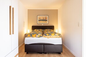A narrow room with a bed featuring a floral bedspread, a headboard, and bedside lamps. - Apartment 'Großes Mehr' with Wi-Fi (Südbrookmerland)