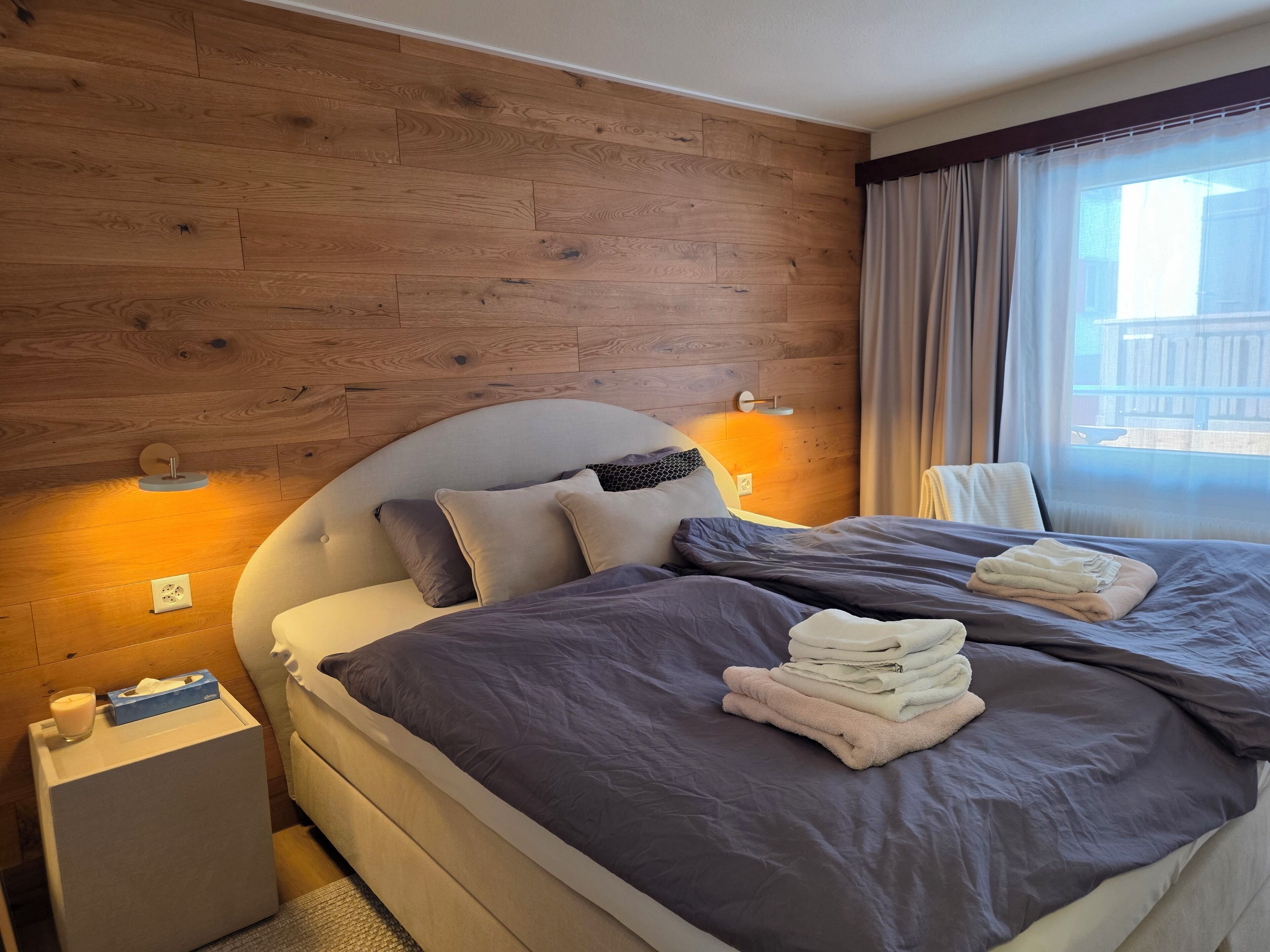 A hotel room with a wooden panel wall, a bed with grey bedding, a bedside table with a candle and tissues, and a window with curtains.