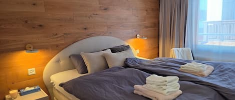 Habitación de hotel con pared de paneles de madera, cama con ropa de cama gris, mesita de noche con una vela y pañuelos, y ventana con cortinas.