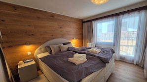 Un dormitorio moderno con una cama grande, paredes de paneles de madera, una mesita de noche y vistas al exterior por una ventana.