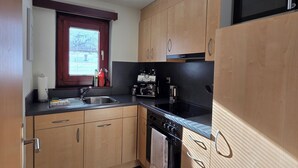 Una cocina compacta con armarios de madera, encimera negra, microondas integrado y una ventana con persianas.