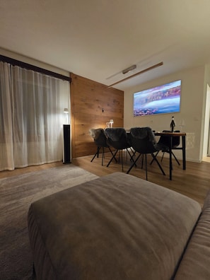 A modern bedroom with a large bed, a wooden accent wall, a dining area with a table and chairs, and a TV displaying a scenic landscape.