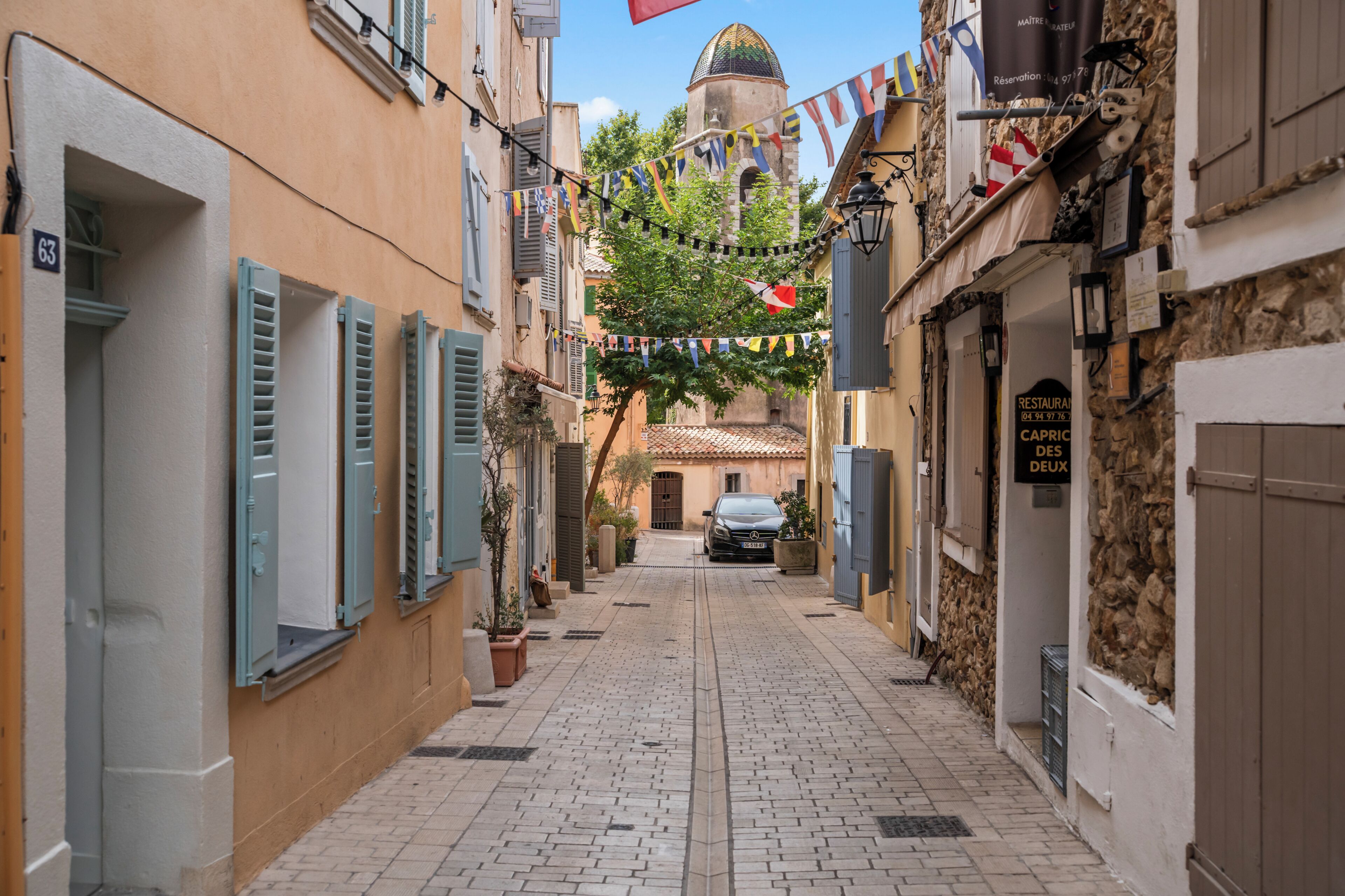 A narrow cobblestone street with buildings on either side, some with shutters, and a car parked in the distance.