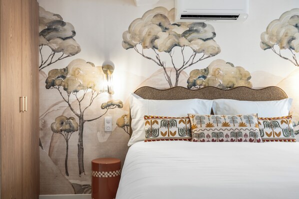 A bedroom with a bed, pillows, a wall-mounted air conditioner, and a wall with a nature-themed mural.