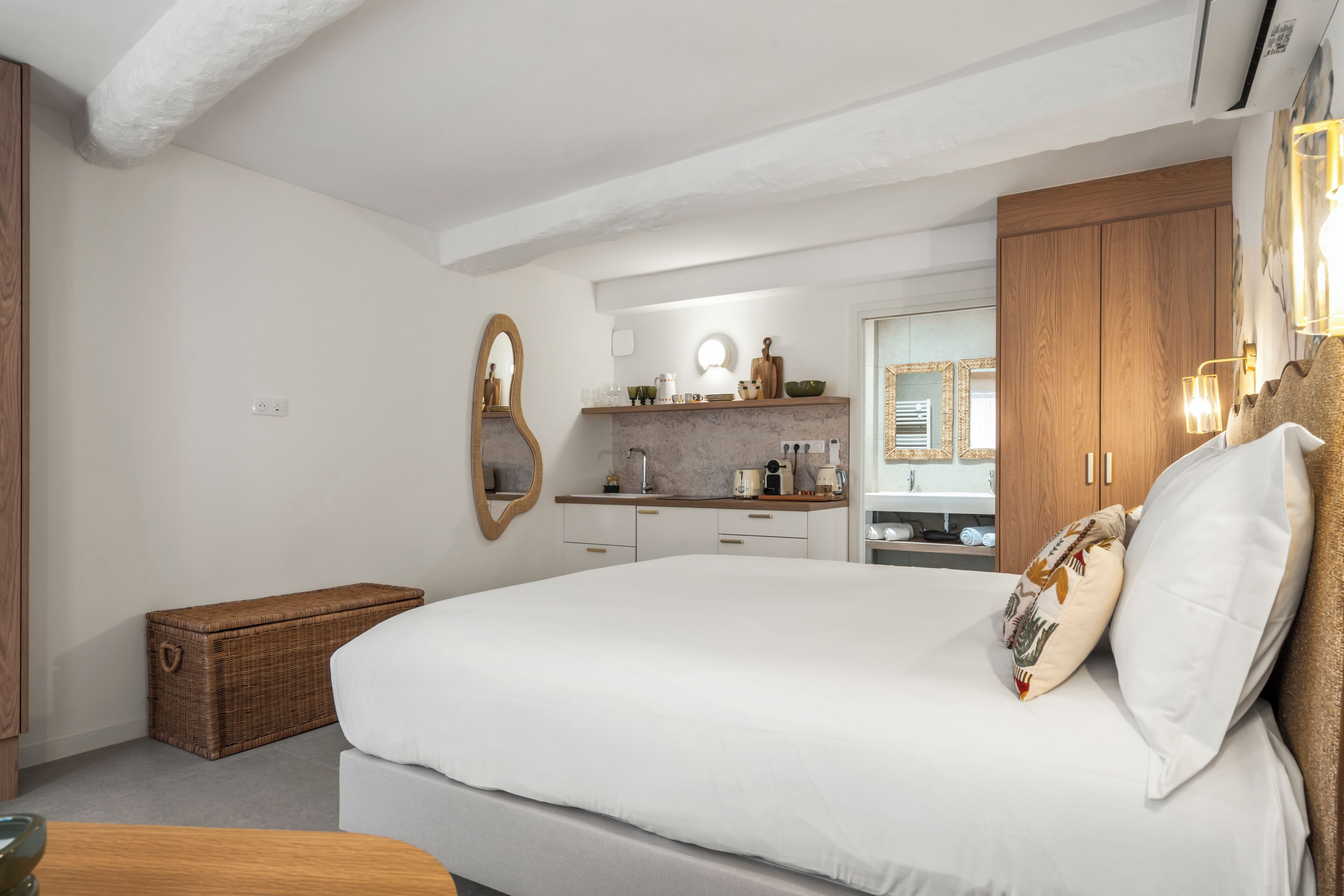 A bedroom with a large bed, a wooden wardrobe, a wicker chest, and a bathroom with a mirror and sink.