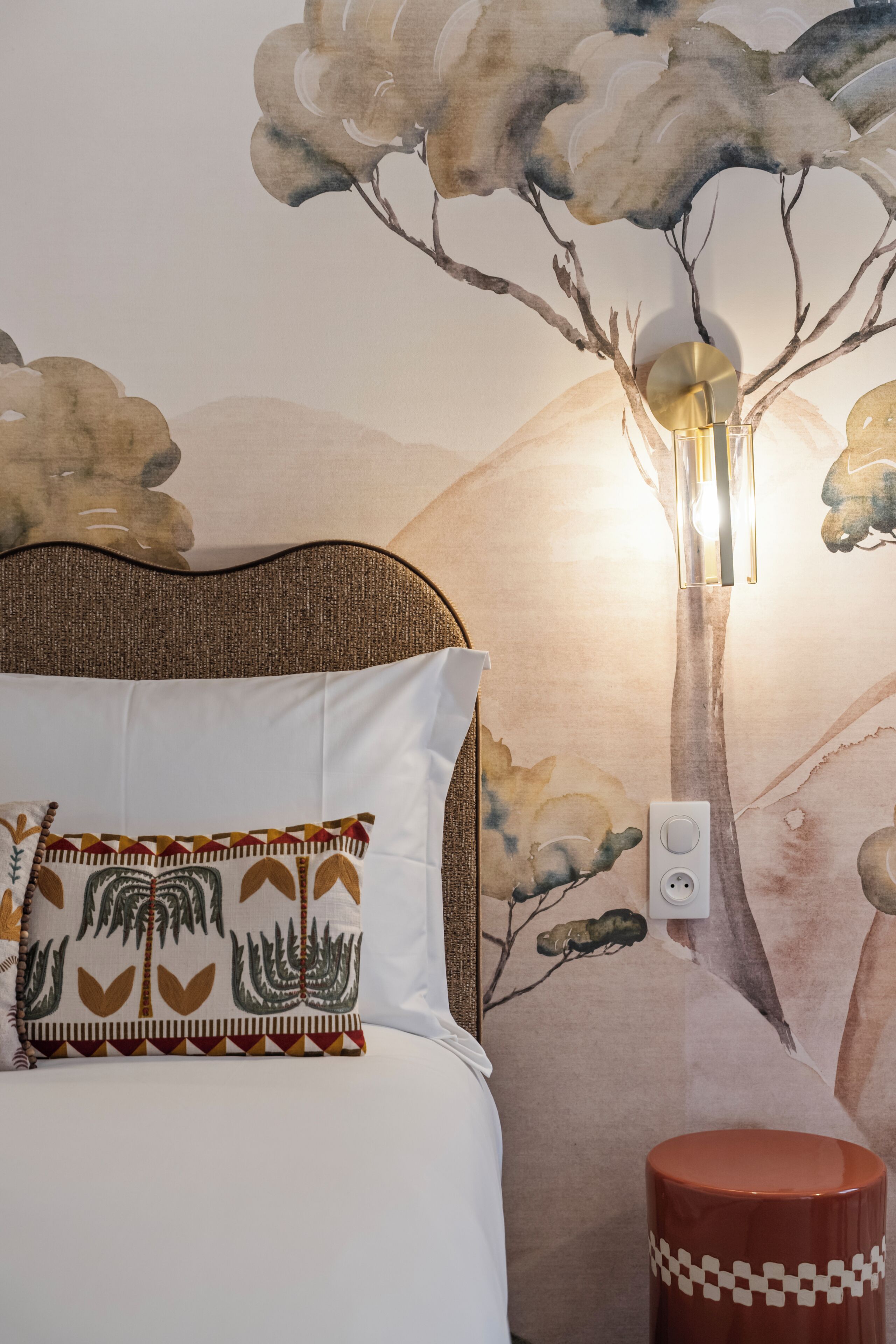 A bed with a patterned pillow, a wall-mounted lamp, and a partial view of a mural.