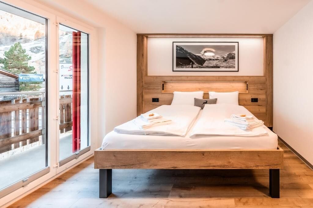 A bedroom with a wooden bed, white bedding, a framed picture on the wall, and a view through a glass door.