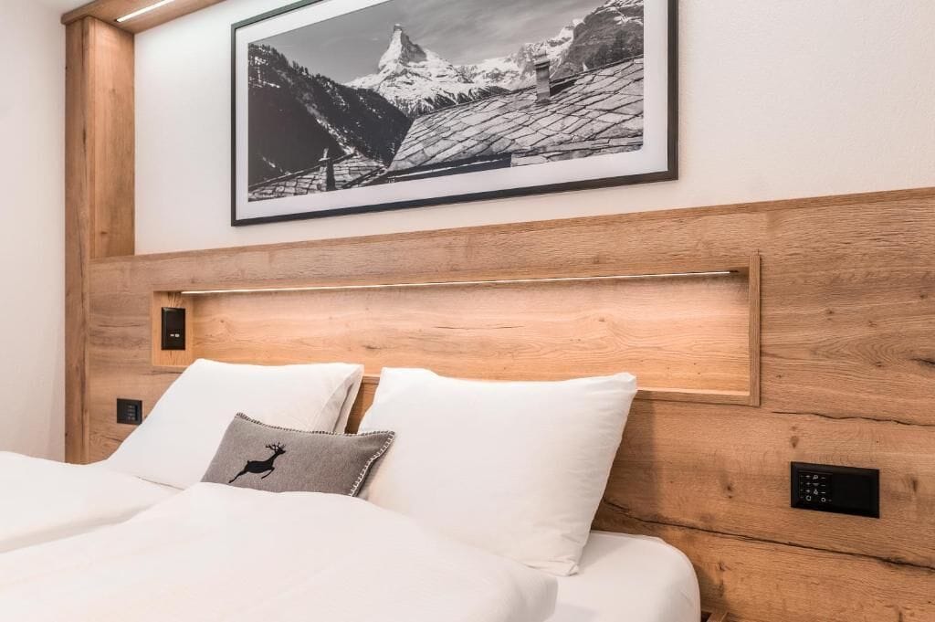 Une chambre d’hôtel avec une tête de lit en bois, un lit recouvert de draps blancs, un oreiller orné d’un motif de cerf et, au mur, un tableau représentant un paysage de montagne.