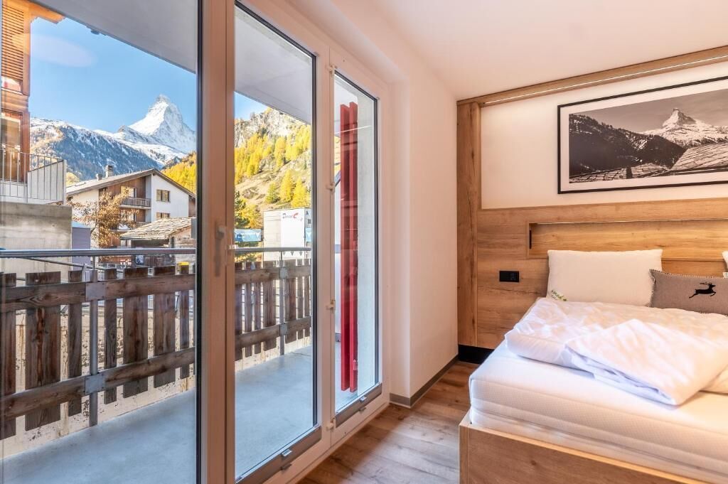 A bedroom with a wooden headboard, a bed with white bedding, a large window with a view of mountains, and a framed picture on the wall.