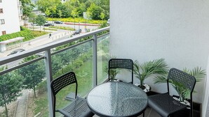 En balkong med ett runt glasbord och fyra stolar, med utsikt över en stadsmiljö med byggnader och grönska.