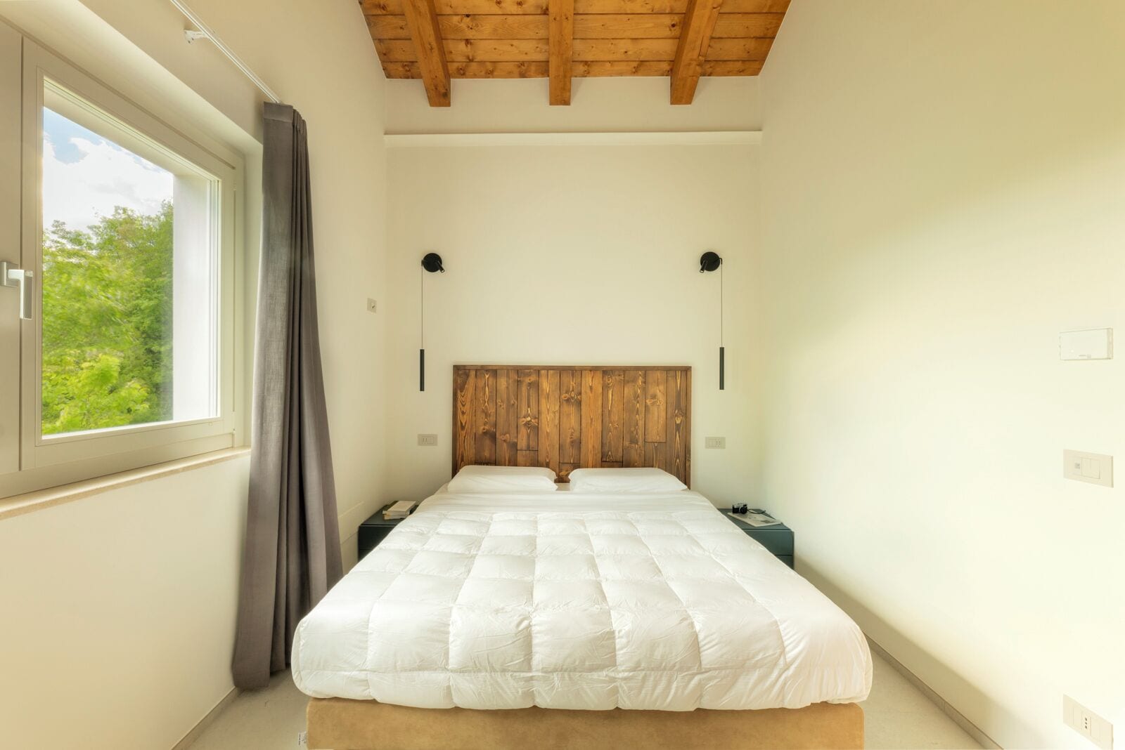 Une chambre à coucher avec une tête de lit en bois, une grande fenêtre avec des rideaux, un lit avec une literie blanche et des lampes de chevet.