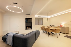 A modern living room with a gray sofa, a dining area with a wooden table and chairs, and a kitchen area with a white cabinet and a window. - VILLA SERRA - Sibillini Retreat with sauna and hot tub (Marche)