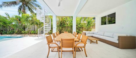 A covered outdoor dining area with a pool, wooden furniture, and lush greenery.