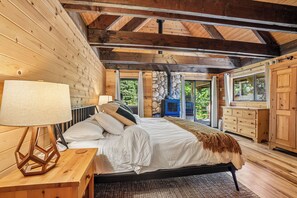 A bedroom with a bed, a nightstand, a lamp, a stone wall, and a view of greenery outside.