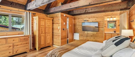 A wooden cabin interior with a bed, dresser, and a large window.