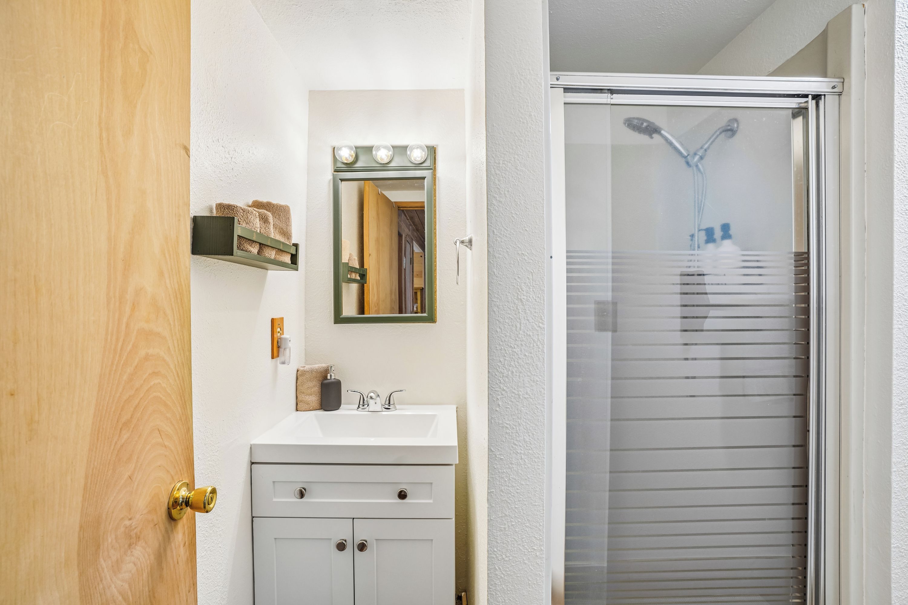 Une salle de bain avec un lavabo blanc, un miroir avec éclairage intégré, un porte-serviettes et une douche dotée d’une porte en verre.