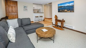 Uma sala de estar com um sofá cinza, uma mesa de centro de madeira e uma pintura de uma paisagem costeira.