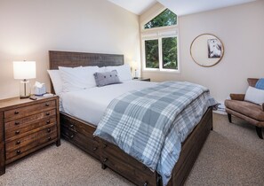 Une chambre avec un lit en bois, une table de chevet avec des tiroirs, une lampe, une fenêtre donnant sur un paysage verdoyant et un miroir rond accroché au mur.