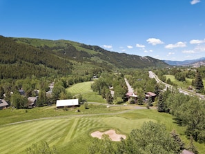 Ein Golfplatz mit Clubhaus, umgeben von bewaldeten Hügeln und blauem Himmel.