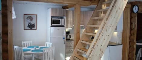 Une cuisine compacte avec des meubles blancs, un four à micro-ondes et un réfrigérateur. Un escalier en bois mène à une zone aménagée en chambre à coucher.