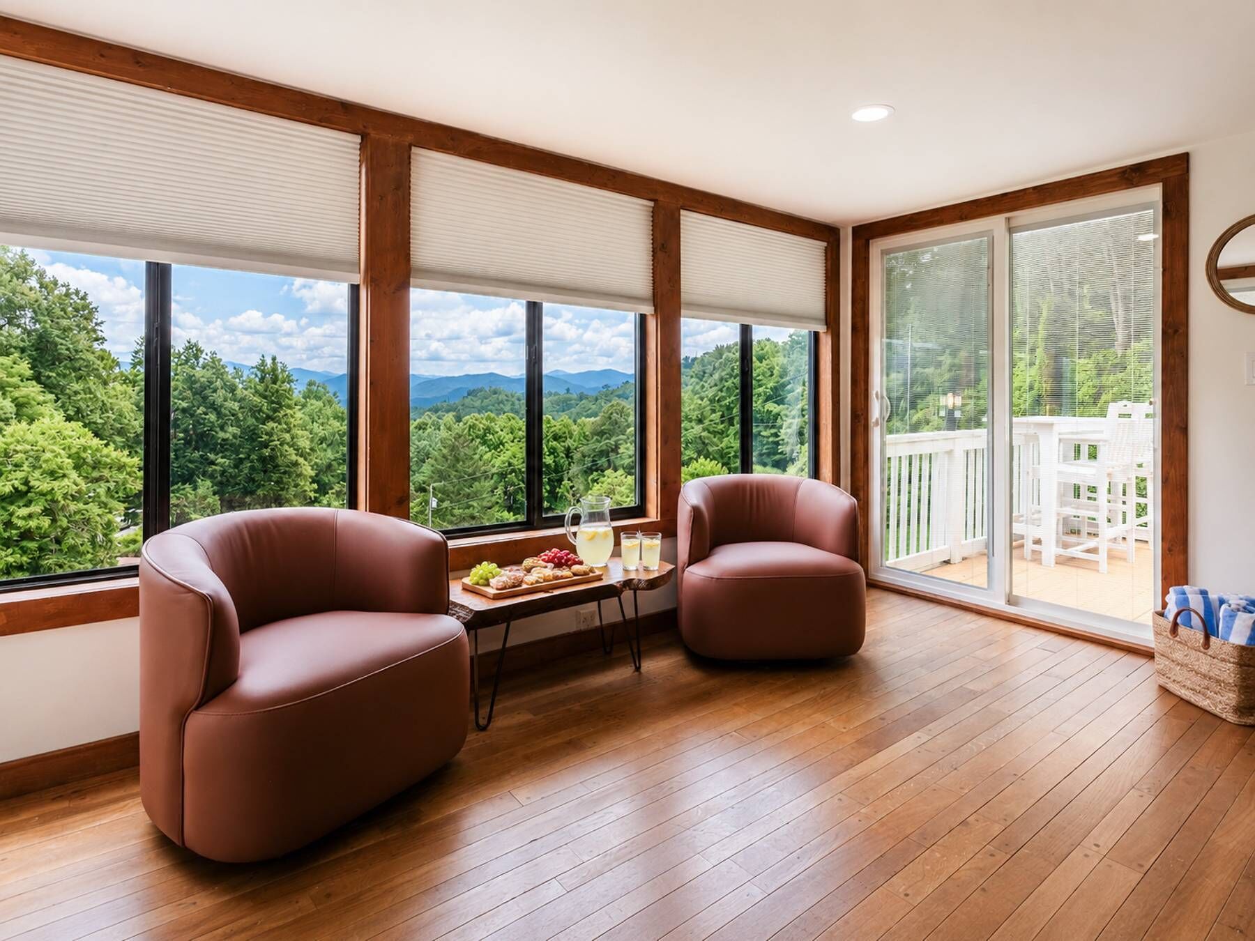 A spacious room with large windows offering a view of a forest and mountains, two leather armchairs, a small table with a pitcher and glasses, and a wooden floor.
