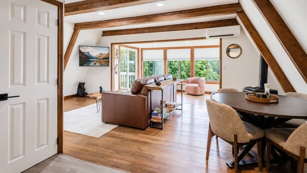 Ein Dachzimmer mit Holzbalken, einem Sofa, einem Esstisch und Blick auf Grün durch große Fenster.