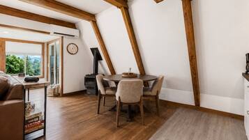 Een knus vertrek met houten plafond, een ronde tafel met stoelen, een open haard en een raam met uitzicht op groen.