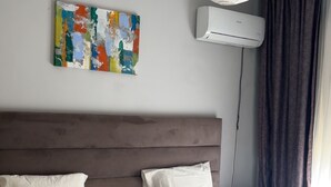 A bedroom with two beds, a ceiling fan, an air conditioning unit, a colorful abstract painting, and a window with curtains.