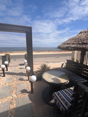 An outdoor seating area with a view of the beach, stone flooring, and a thatched-roof structure.