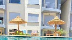 A swimming pool with lounge chairs and thatched umbrellas, surrounded by a multi-story building with balconies.