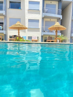 A swimming pool with lounge chairs and thatched umbrellas, surrounded by a multi-story building with balconies.