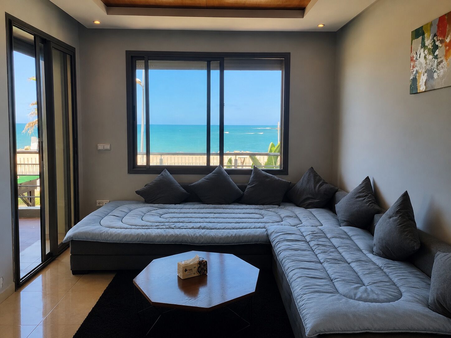 Ein modernes Wohnzimmer mit großem Fenster zum Meer, einem Ecksofa und einem kleinen Tisch.