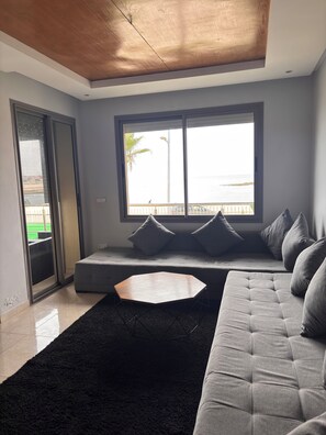 A modern living room with a sofa, a coffee table, and a large window offering a view of the sea.