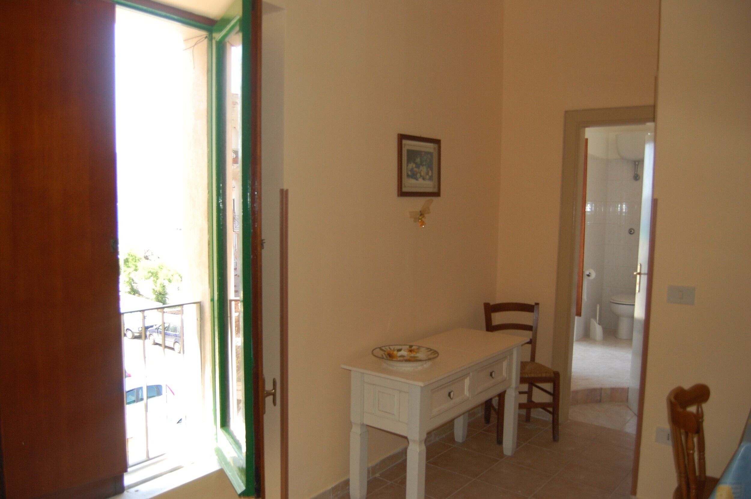 Una stanza con un tavolo e sedie bianche, una porta che dà su un balcone e un quadro appeso al muro.