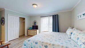 Ein Schlafzimmer mit Bett, Kommode, Fernseher, einer Tür und einem fensterbepflanzten Fenster.