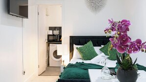 A bedroom with a bed, green pillows, a potted orchid, a glass vase, and a wall-mounted TV. - Studio Sleeps 2 With Parking (Thornton Heath)