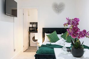 A bedroom with a bed, green pillows, a potted orchid, a glass vase, and a wall-mounted TV. - Studio Sleeps 2 With Parking (Thornton Heath)